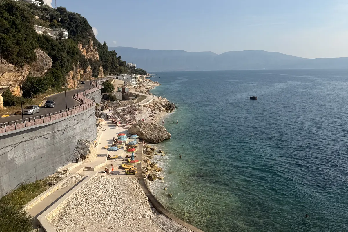 La strada costiera della Riviera albanese sopra una piccola spiaggia di ciottoli con ombrelloni e acqua turchese vicino a Valona.