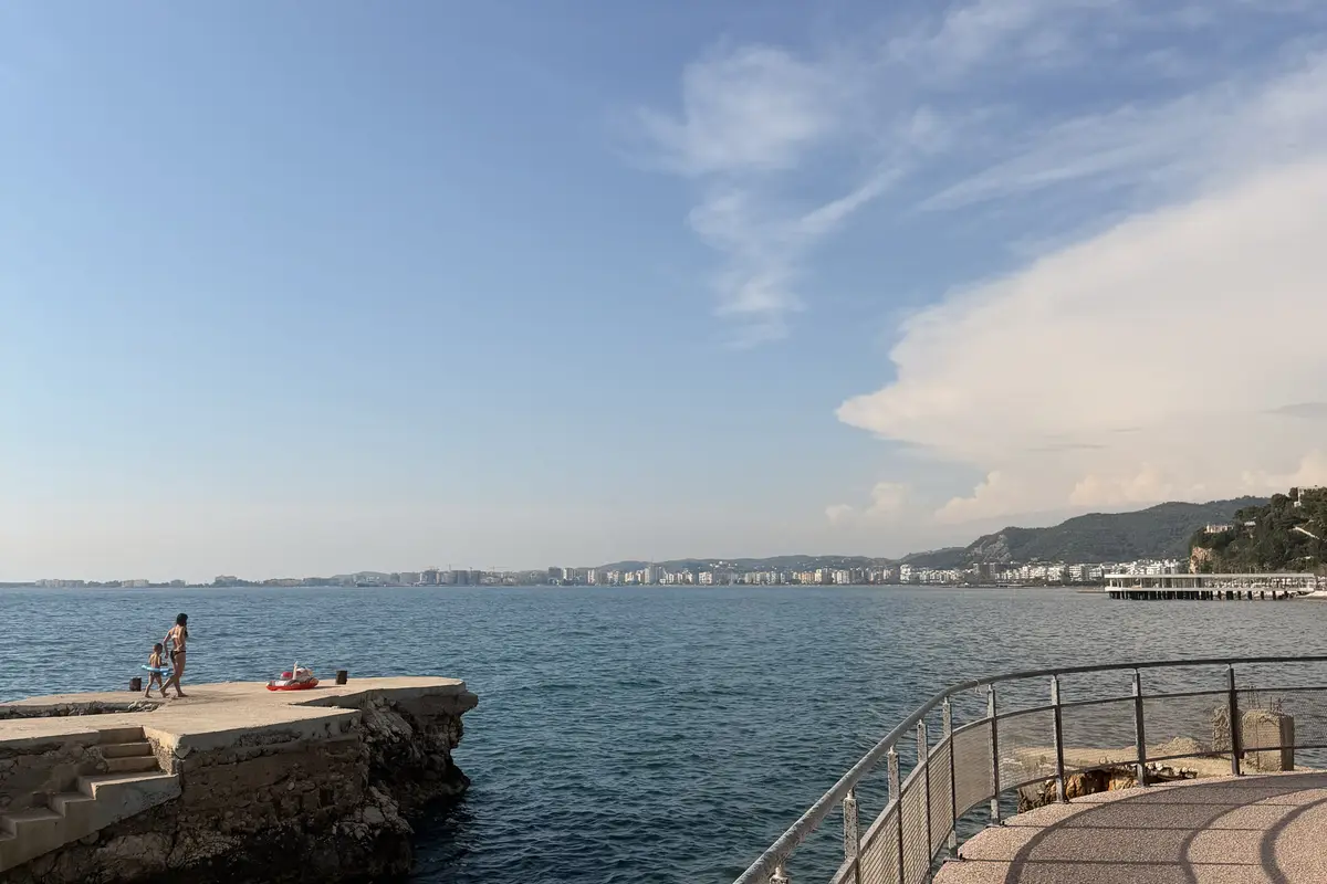 Il lungomare di Valona, in Albania, con una mamma e un bambino che passeggiano su un percorso con ringhiera sopra la baia tranquilla.