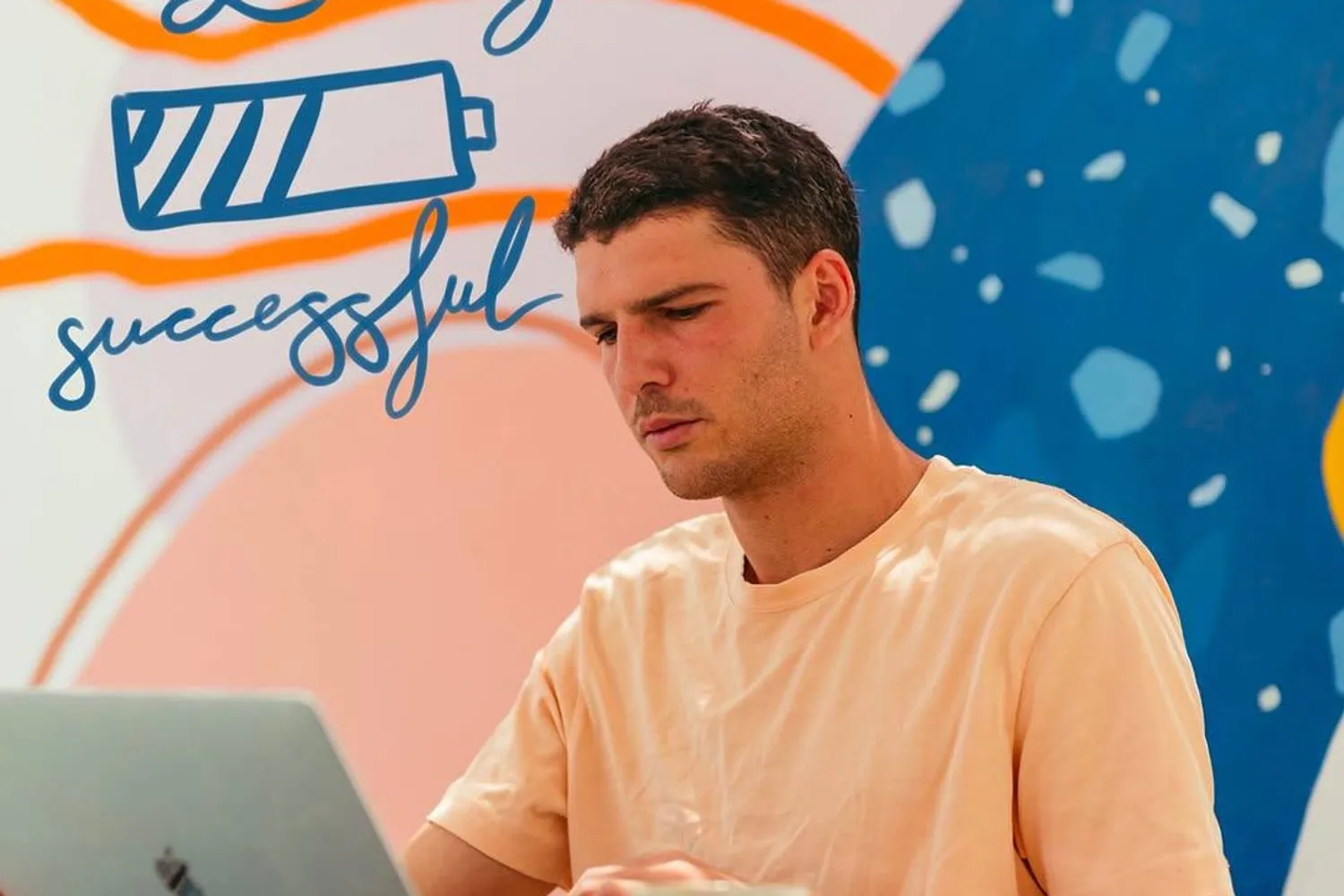 Un ragazzo lavora al portatile con un caffe davanti a un murale colorato di coworking con scritto "Loading successful".